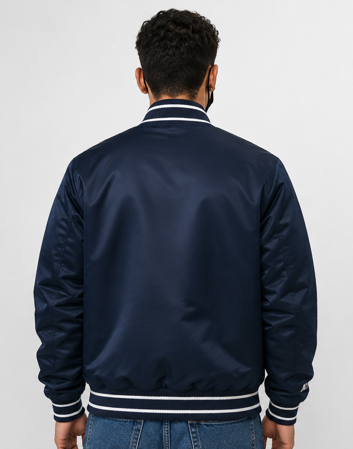 A man viewed from behind wearing a navy blue satin bomber jacket with white striped ribbing on the collar, cuffs, and hem, paired with blue jeans against a light gray background.