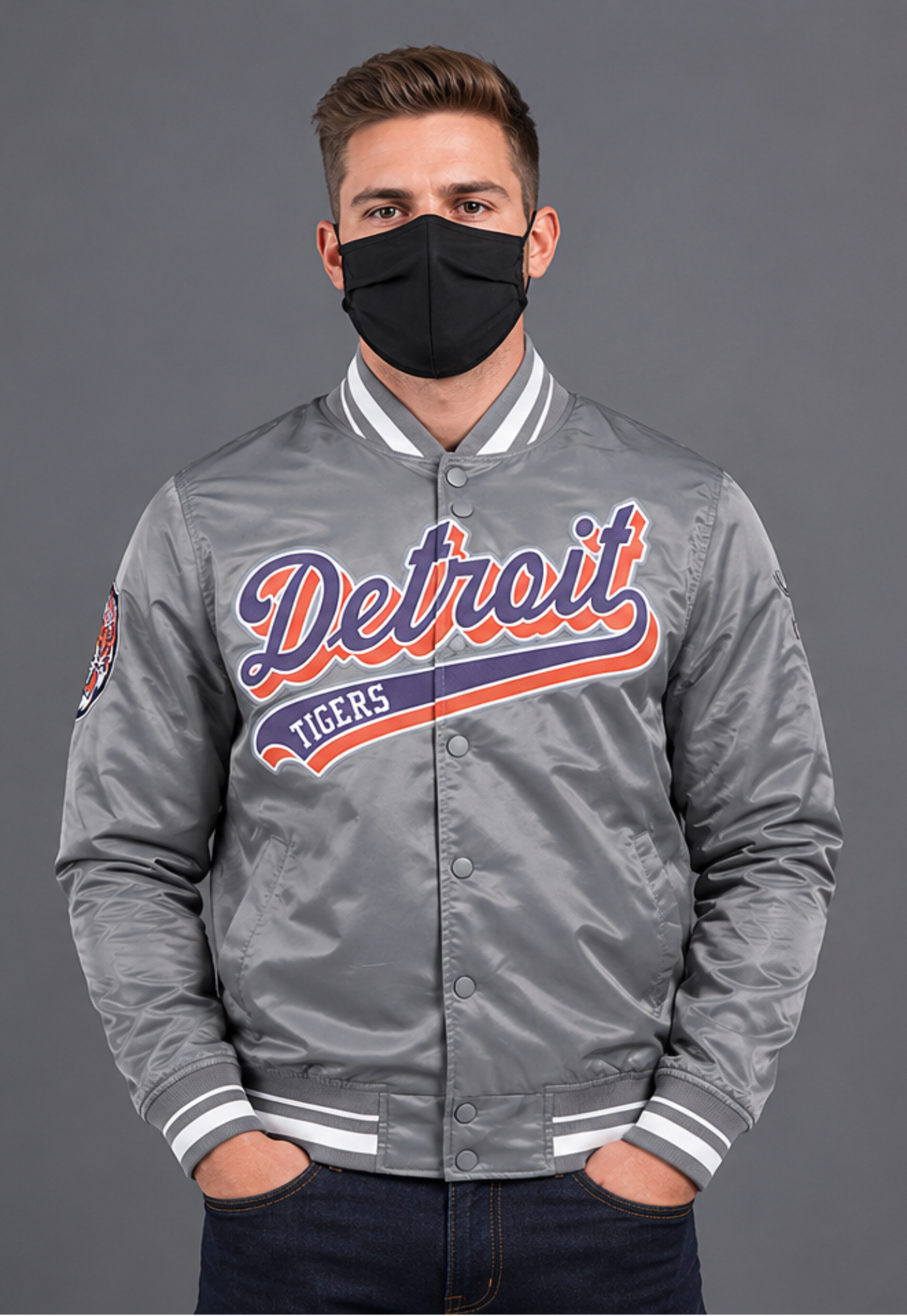 A man wearing a gray satin Detroit Tigers varsity jacket with a black face mask, standing against a solid gray studio background.