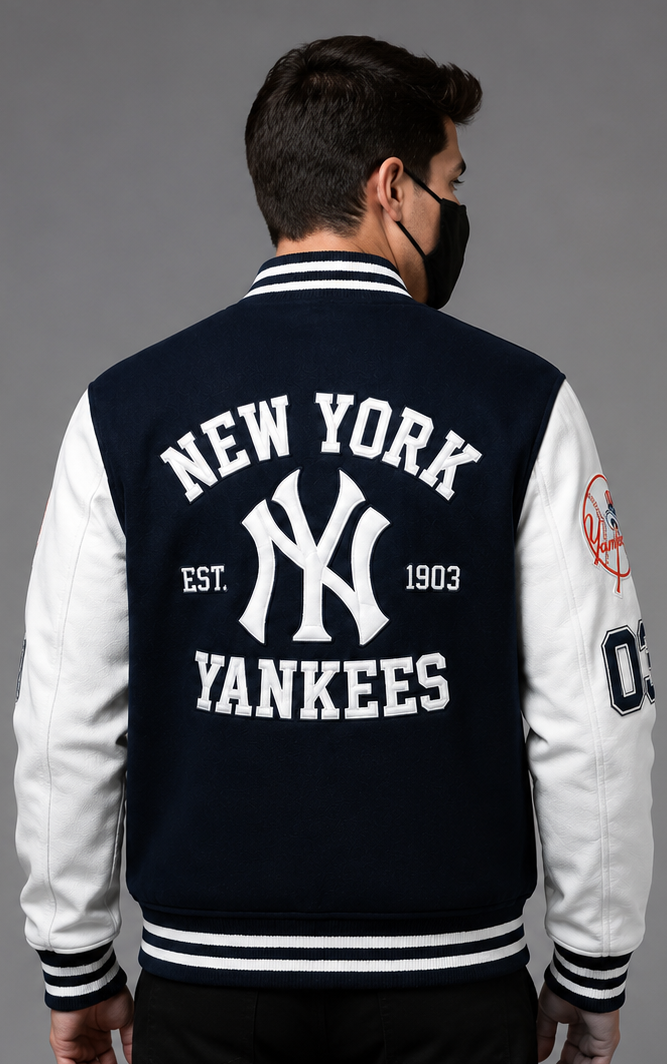 A man viewed from the back wearing a navy and white New York Yankees varsity jacket with a black face mask, set against a solid gray studio background.