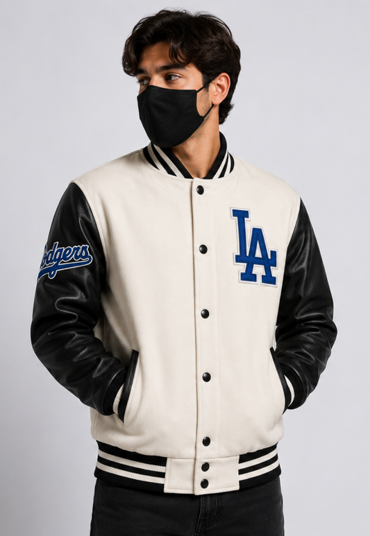 A man wearing a cream and black varsity jacket with blue 'LA' and 'Dodgers' graphics, paired with a black face mask, against a light gray studio background.
