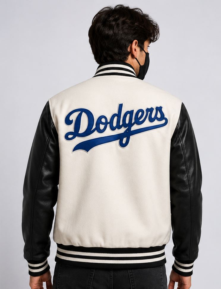 A man viewed from behind wearing a cream and black varsity jacket with 'Dodgers' embroidered in blue on the back, set against a plain light gray studio background.
