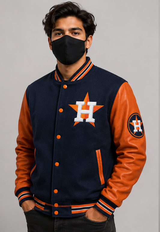A man wearing a navy and orange Houston Astros varsity jacket with a black face mask, standing against a solid gray studio background.
