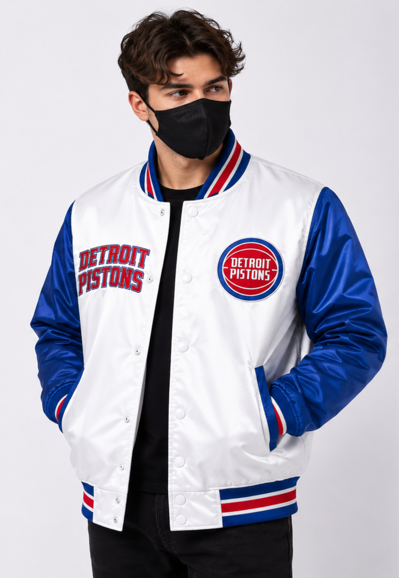 A man wearing a Detroit Pistons varsity jacket with a white satin body, royal blue sleeves, and red and white striped ribbed collar, cuffs, and hem. He is wearing a black face mask and a black shirt underneath, photographed against a light gray studio background.