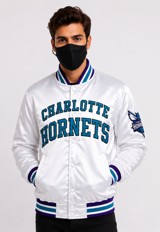 A man wearing a white Charlotte Hornets satin bomber jacket with blue and purple ribbed trim, a black face mask, and black pants, photographed against a light gray studio background.