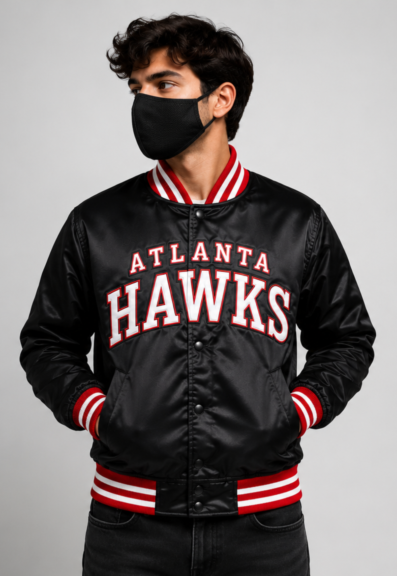 A man wearing a black satin Atlanta Hawks varsity jacket with red and white striped ribbed collar, cuffs, and hem, paired with a black face mask, against a solid gray background.