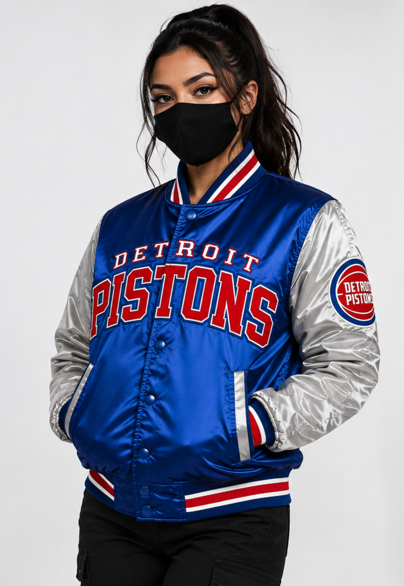 A woman wearing a Detroit Pistons satin varsity jacket with a black face mask, photographed against a light gray studio background.