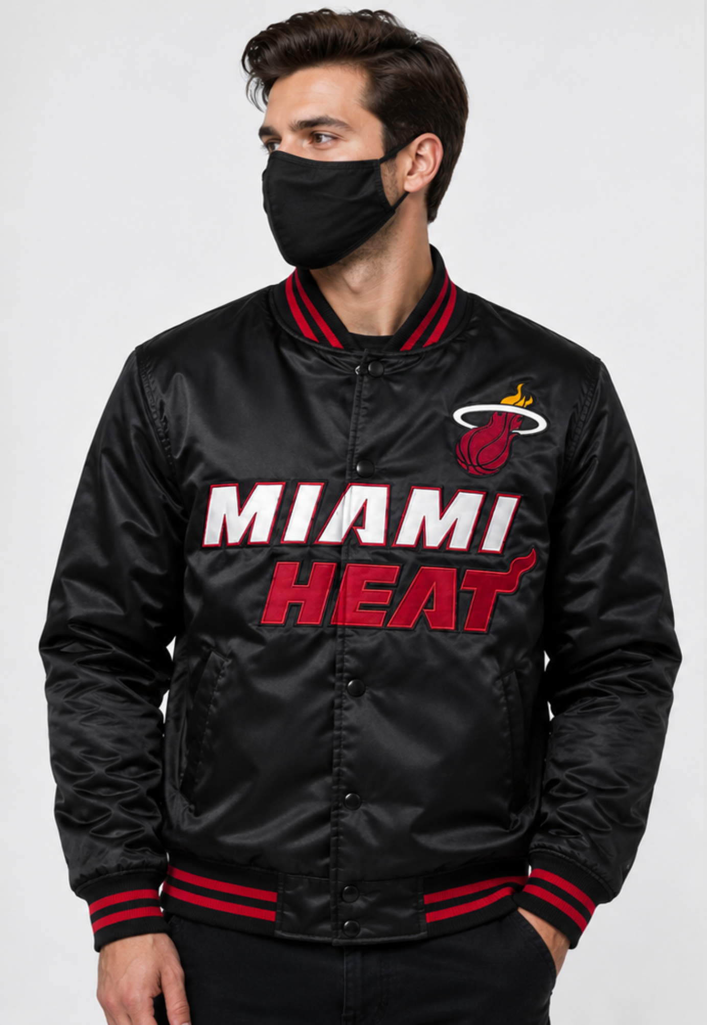 A man wearing a black Miami Heat satin bomber jacket with red and white lettering, paired with a black face mask, against a light gray background.