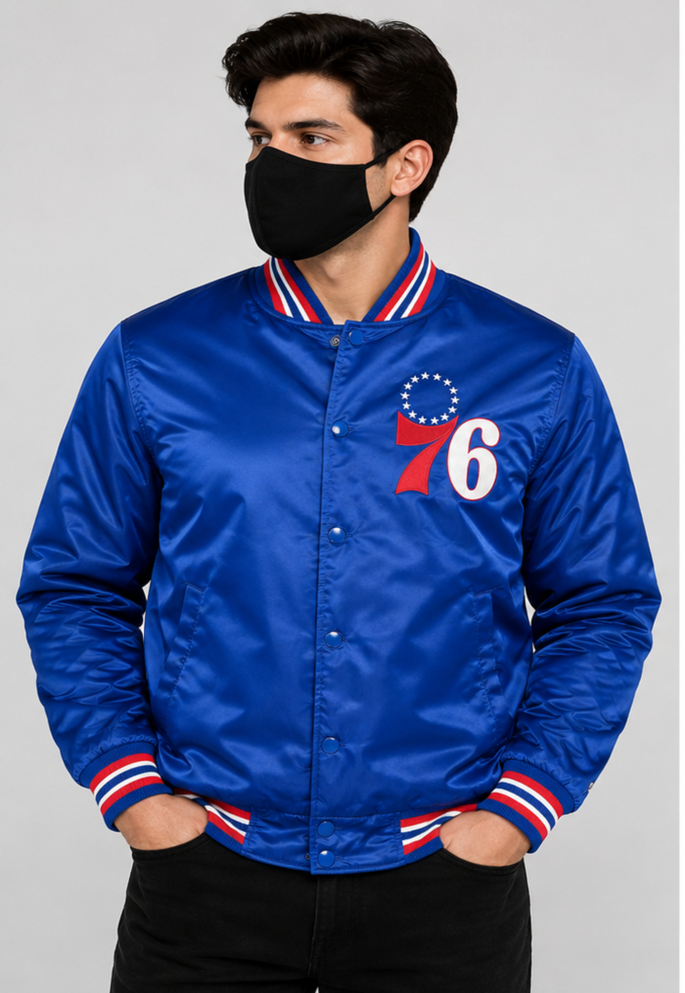 A man wearing a royal blue satin varsity jacket with red and white striped ribbed collar, cuffs, and hem, featuring a '76' chest graphic. He is wearing a black face mask and black pants against a light gray studio background.