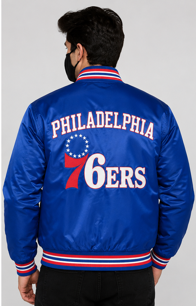A man viewed from the back wearing a royal blue Philadelphia 76ers satin bomber jacket with red and white striped ribbed collar, cuffs, and hem. The back features 'PHILADELPHIA' in white arched letters above a red and white 76ers logo.