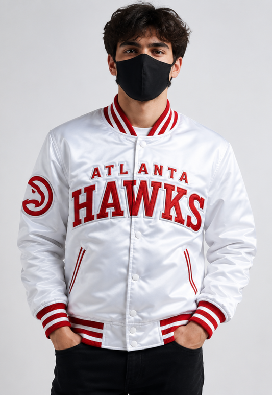 A man wearing a white Atlanta Hawks varsity jacket with red lettering and striped ribbed collar, cuffs, and hem, paired with a black face mask and black pants, against a light gray studio background.