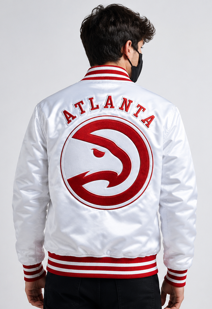 A man viewed from behind wearing a white Atlanta Hawks satin varsity jacket with red and white ribbed trim, featuring the team logo and 'ATLANTA' text on the back.