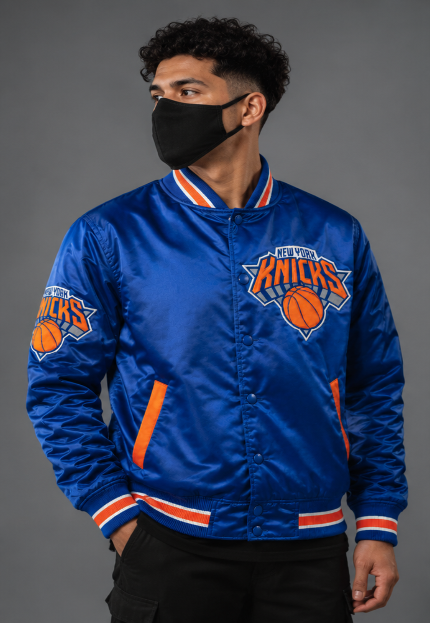 A man wearing a royal blue New York Knicks satin bomber jacket with orange and white trim, a black face mask, and black pants, photographed against a solid gray studio background.