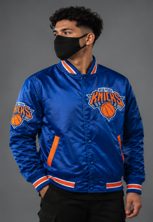 A man wearing a royal blue New York Knicks satin bomber jacket with orange and white trim, a black face mask, and black pants, photographed against a solid gray studio background.