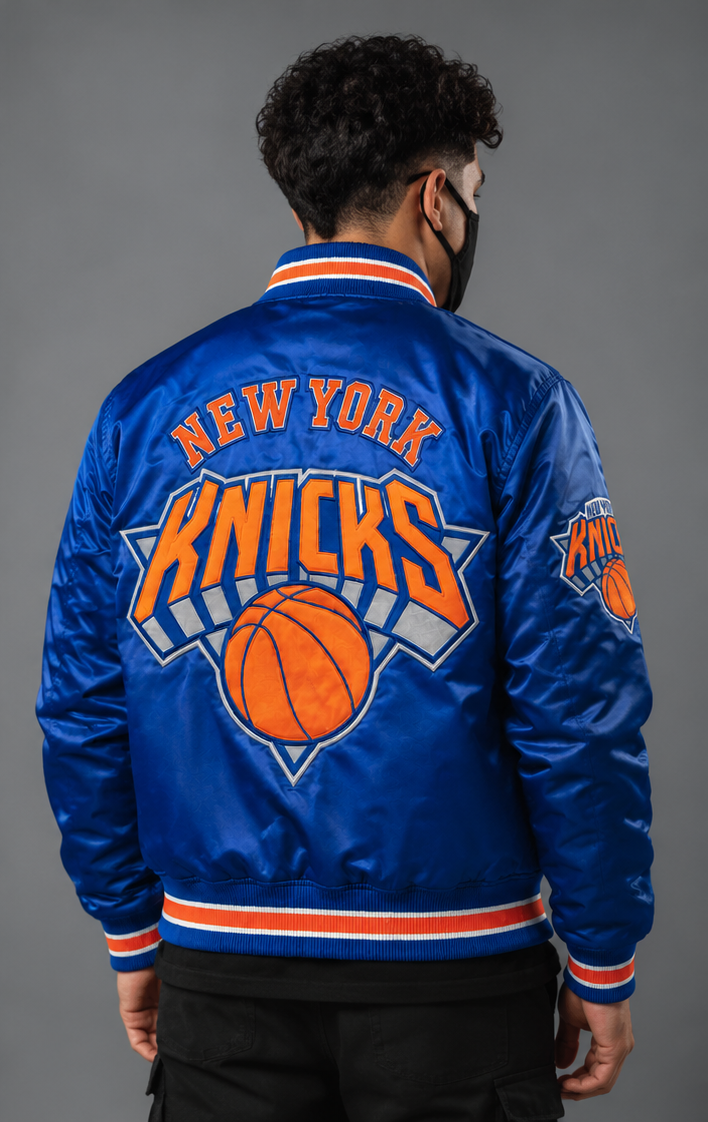 A man viewed from behind wearing a royal blue satin New York Knicks bomber jacket with orange and white lettering and graphics on the back, paired with black pants against a gray studio background.