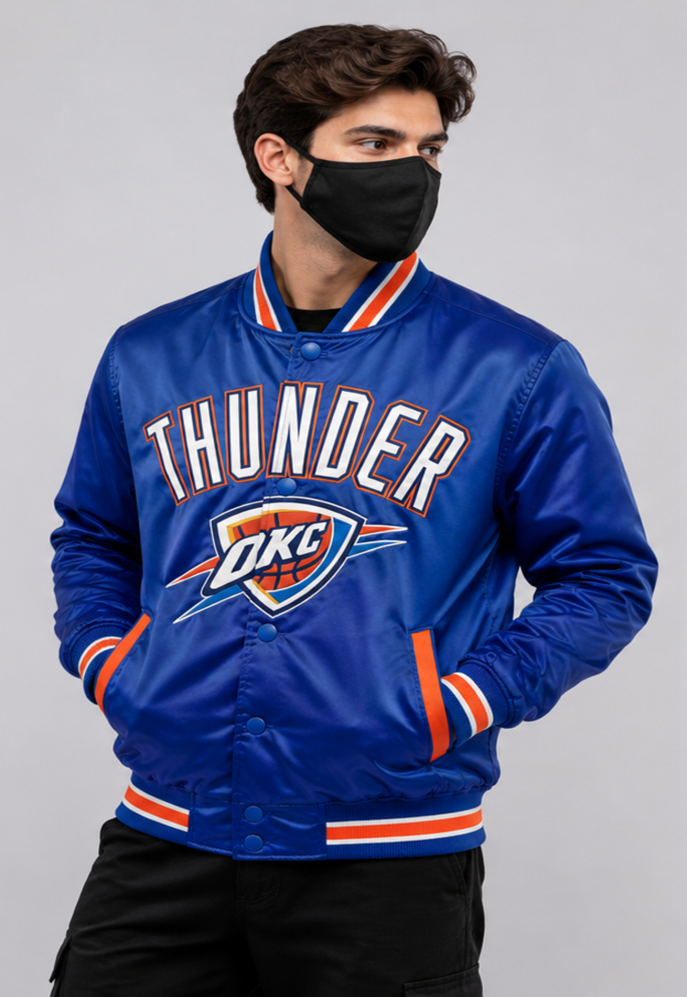 A man wearing a blue Oklahoma City Thunder satin bomber jacket with orange and white trim, a black face mask, and black pants, photographed against a solid gray studio background.