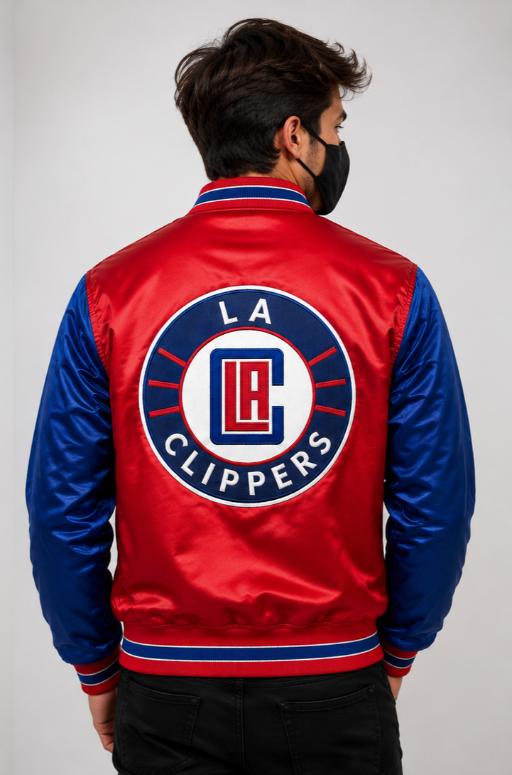 A man viewed from behind wearing a red and blue satin bomber jacket with the Los Angeles Clippers logo on the back, paired with a black face mask and black pants, against a light gray studio background.
