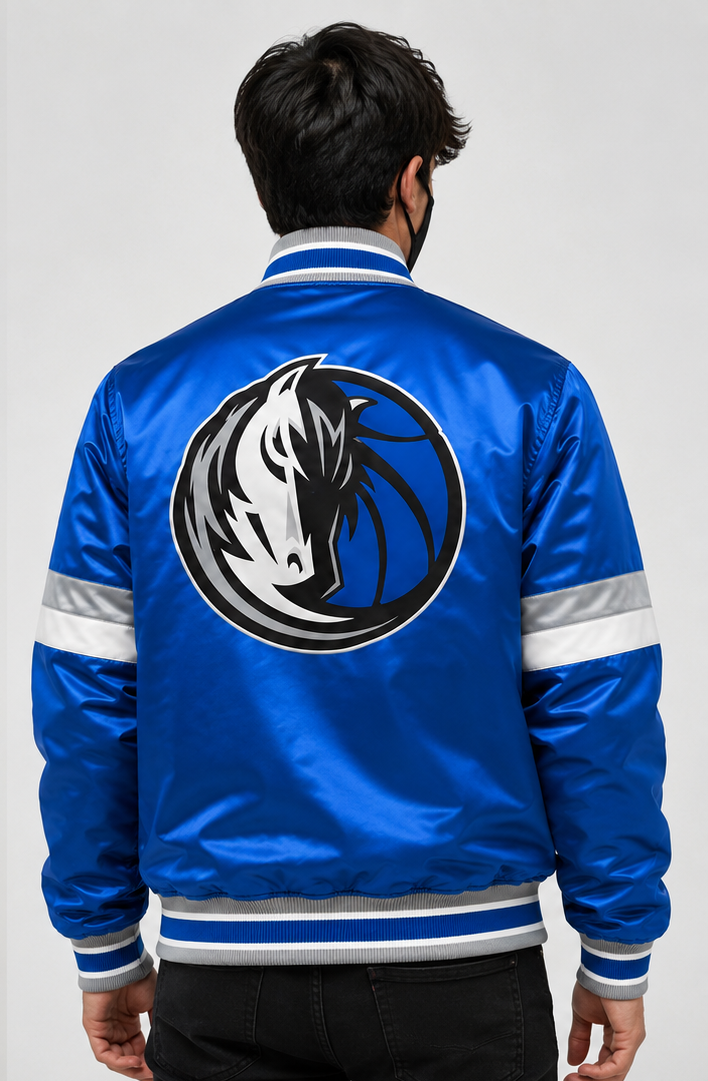 A man viewed from behind wearing a blue satin varsity-style jacket with a large Dallas Mavericks horse logo on the back, silver reflective stripes on the sleeves, and a striped collar, cuffs, and hem.