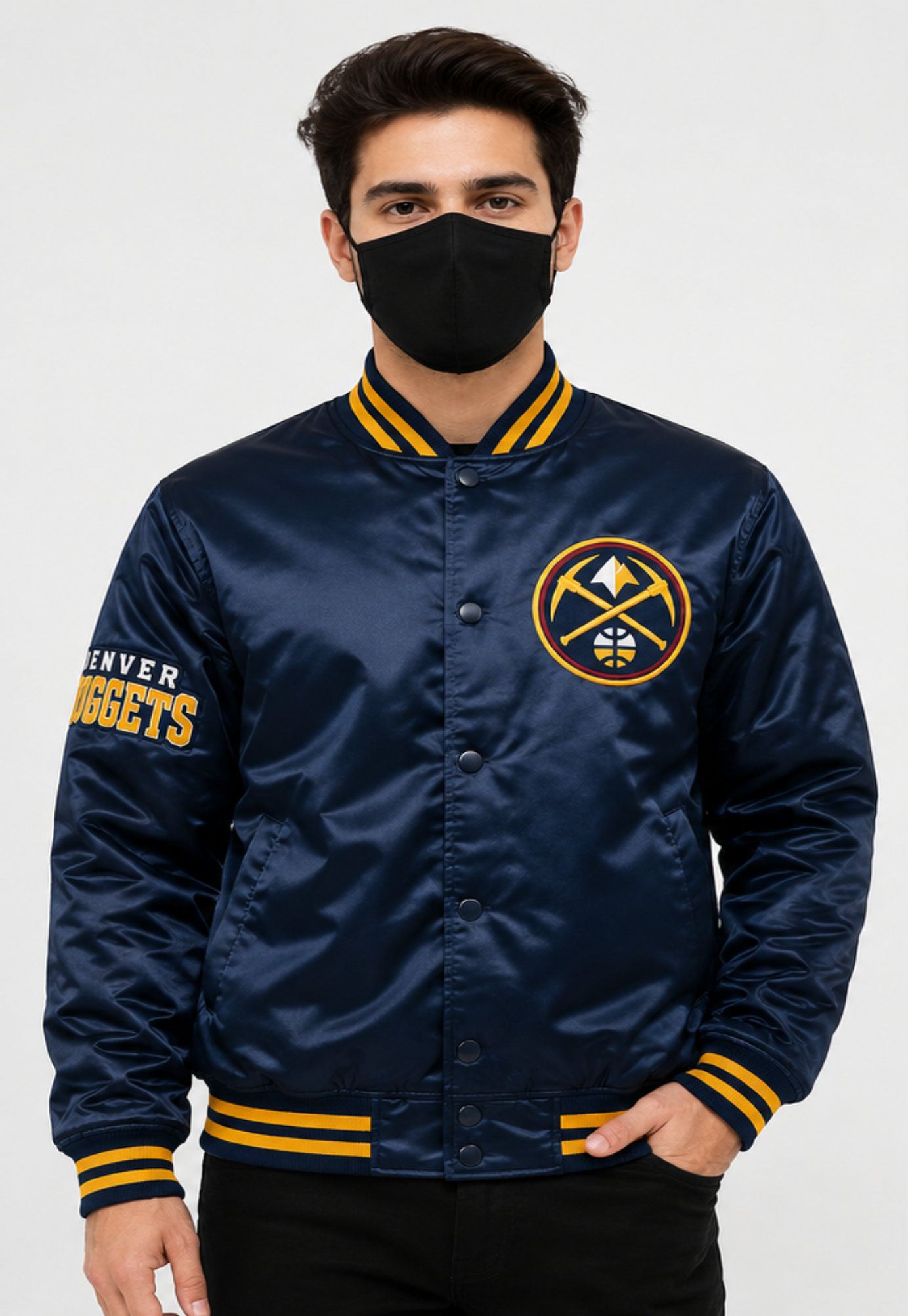 A man wearing a navy blue Denver Nuggets satin bomber jacket with yellow and white accents, paired with a black face mask and black pants, against a light gray studio background.