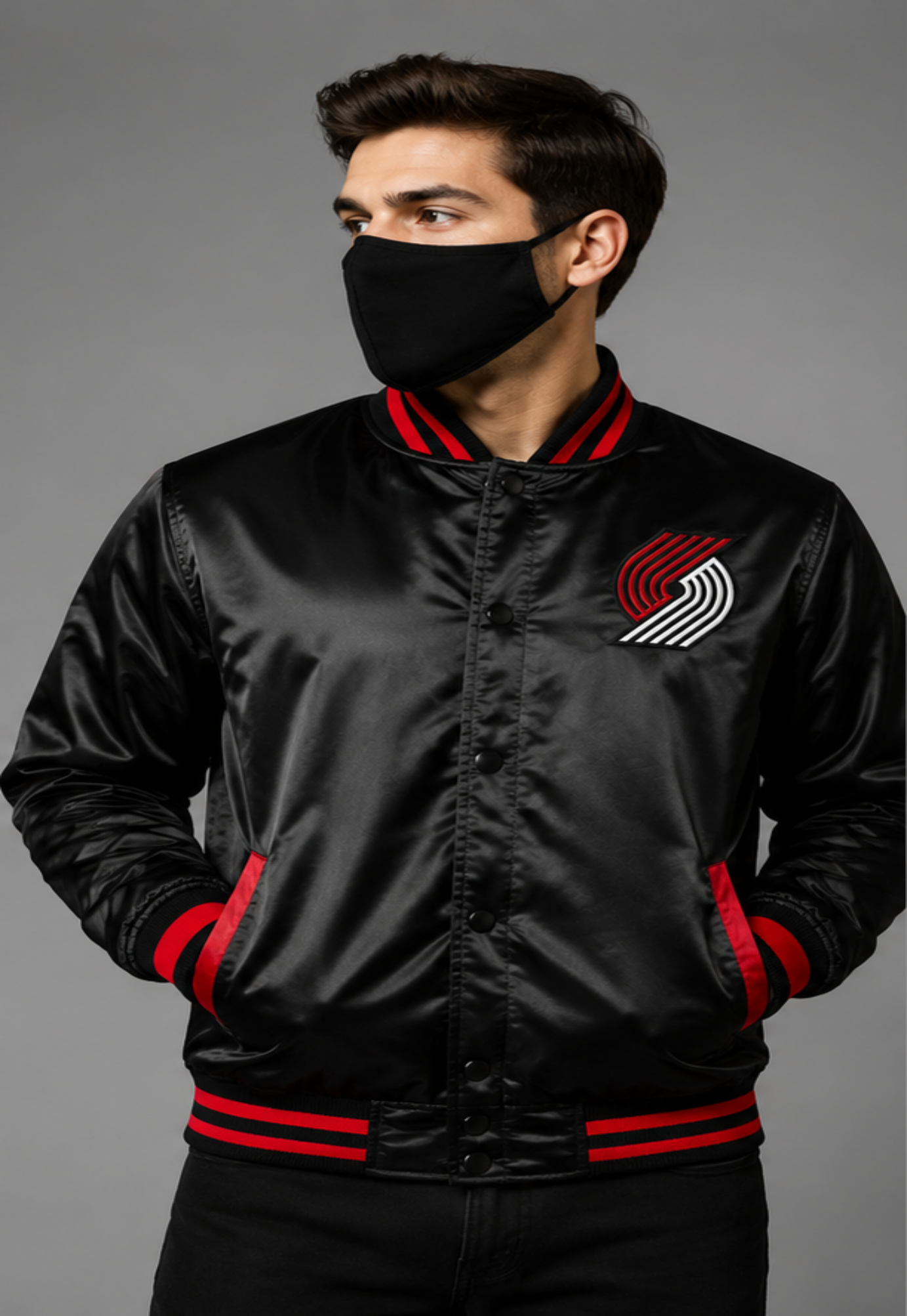 A man wearing a black satin bomber jacket with red and white striped ribbed collar, cuffs, and hem, featuring a red and white Portland Trail Blazers logo on the left chest, set against a solid gray studio background.