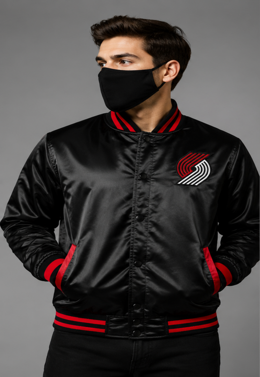 A man wearing a black satin bomber jacket with red and white striped ribbed collar, cuffs, and hem, featuring a red and white Portland Trail Blazers logo on the left chest, set against a solid gray studio background.