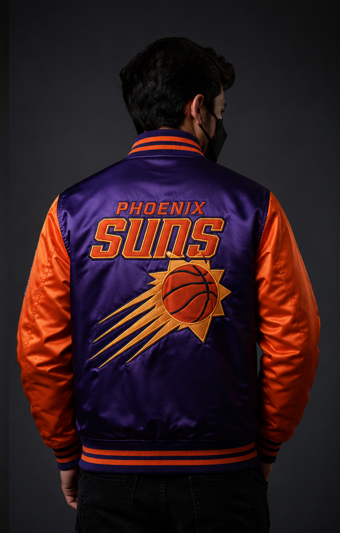A man viewed from behind wearing a Phoenix Suns satin varsity jacket with purple and orange panels and a large basketball logo on the back, against a dark gray studio background.