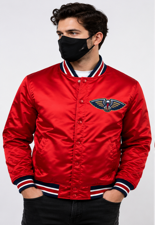 A man wearing a red satin bomber jacket with the New Orleans Pelicans logo on the chest, paired with a black face mask and black pants, against a light gray background.