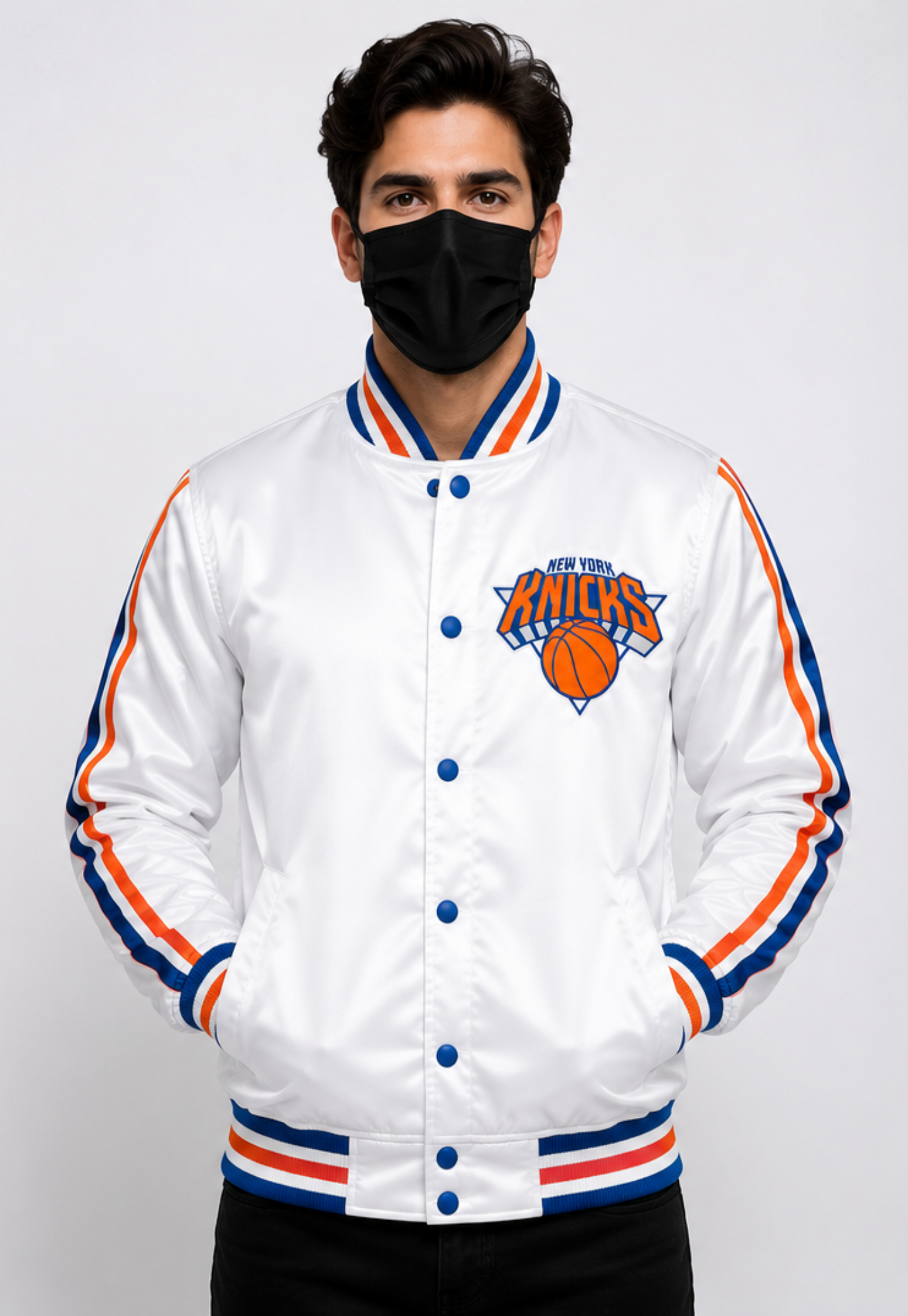 A man wearing a white New York Knicks satin bomber jacket with blue and orange stripes on the collar, cuffs, and hem, paired with a black face mask and black pants, against a light gray background.