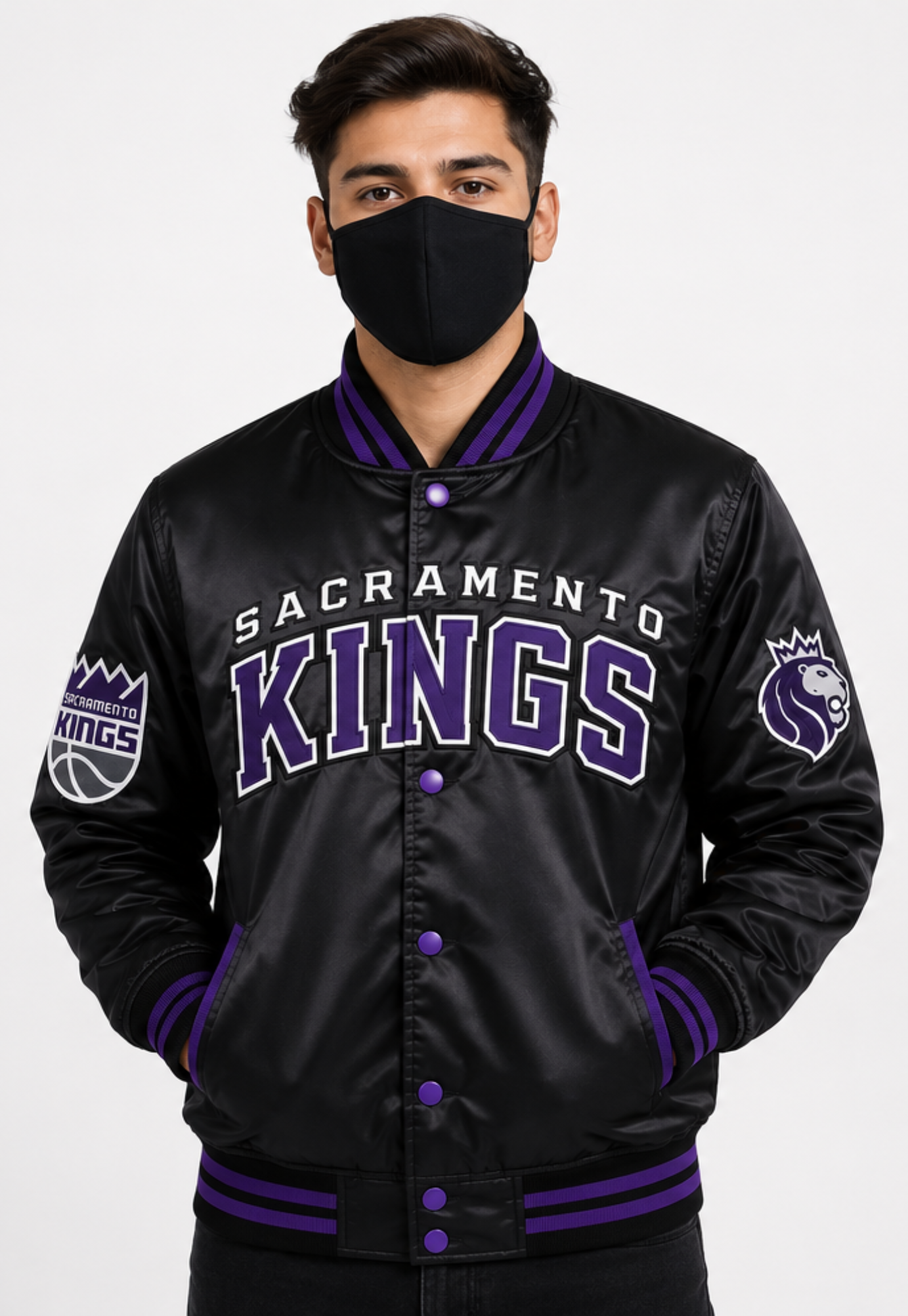 A man wearing a black Sacramento Kings satin varsity jacket with purple and white lettering, a black face mask, and standing against a light gray background.