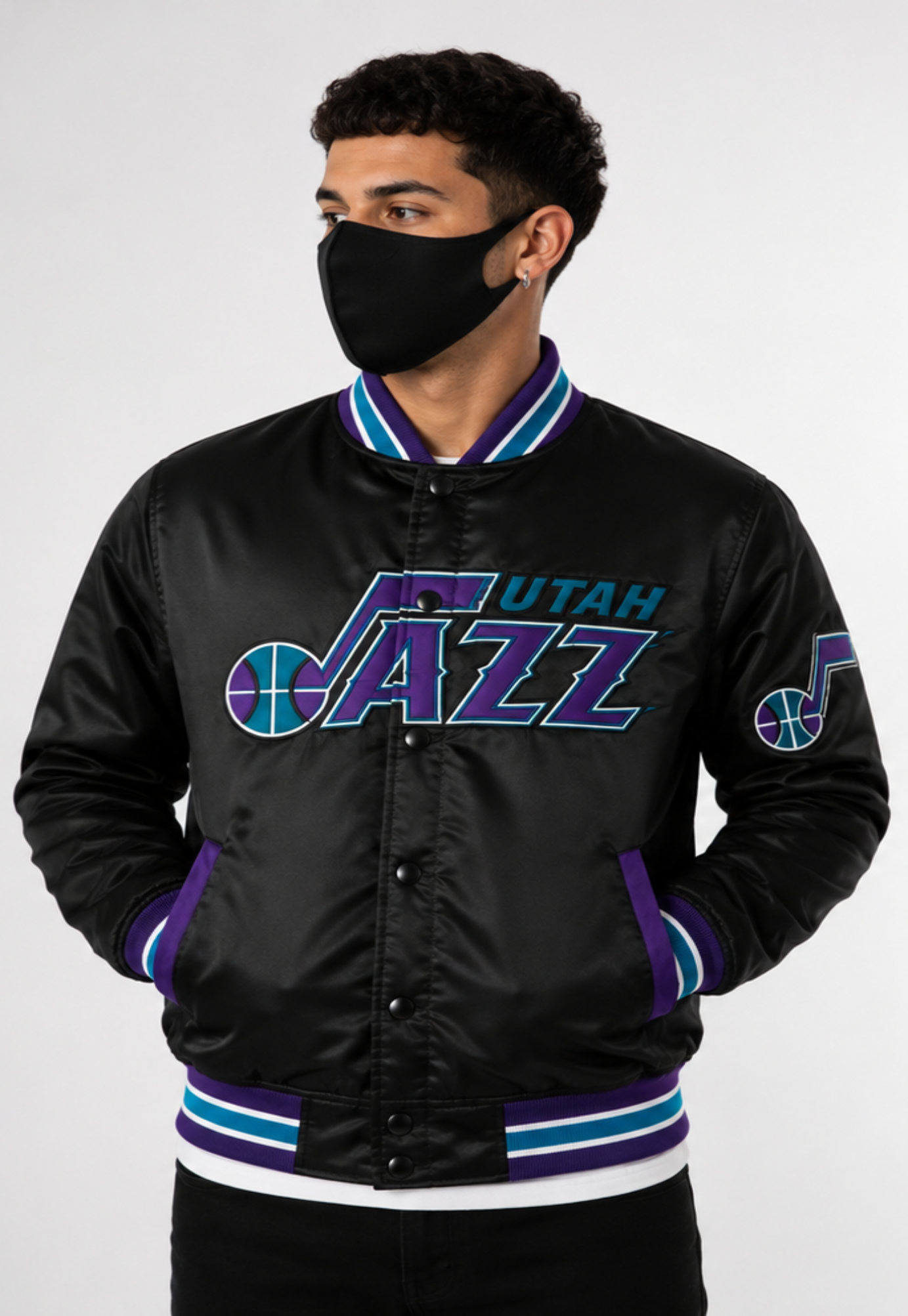 A man wearing a black satin Utah Jazz varsity jacket with purple and teal accents, paired with a black face mask, against a light gray studio background.