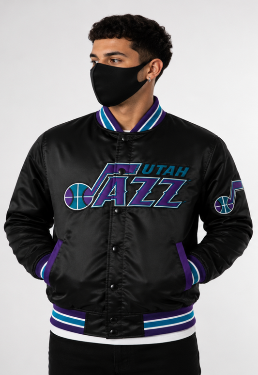 A man wearing a black satin Utah Jazz varsity jacket with purple and teal accents, paired with a black face mask, against a light gray studio background.