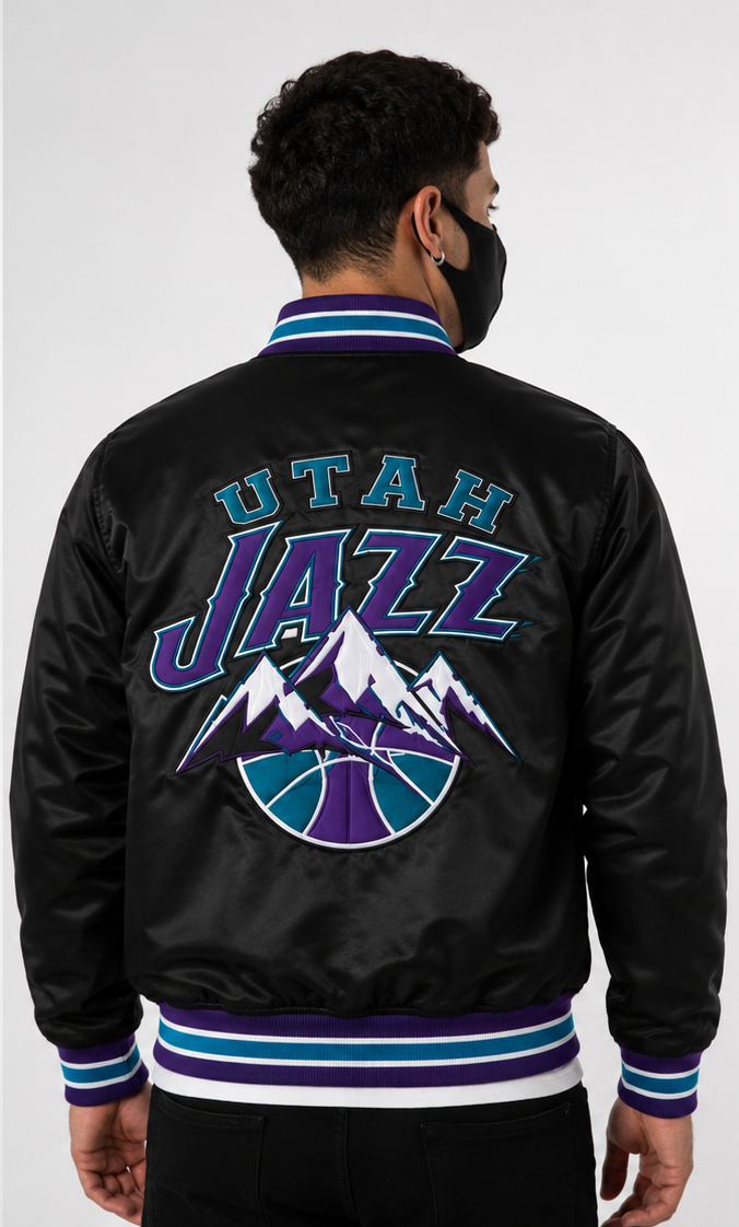 A man viewed from the back wearing a black satin Utah Jazz bomber jacket with a large embroidered team logo on the back and striped ribbed collar, cuffs, and hem.