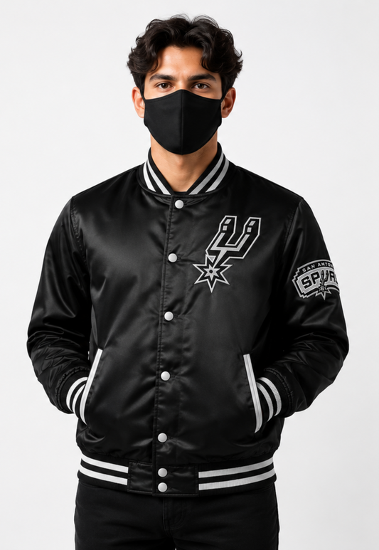 A man wearing a black satin bomber jacket with the San Antonio Spurs logo on the chest and a Spurs patch on the sleeve, paired with a black face mask, against a light gray studio background.