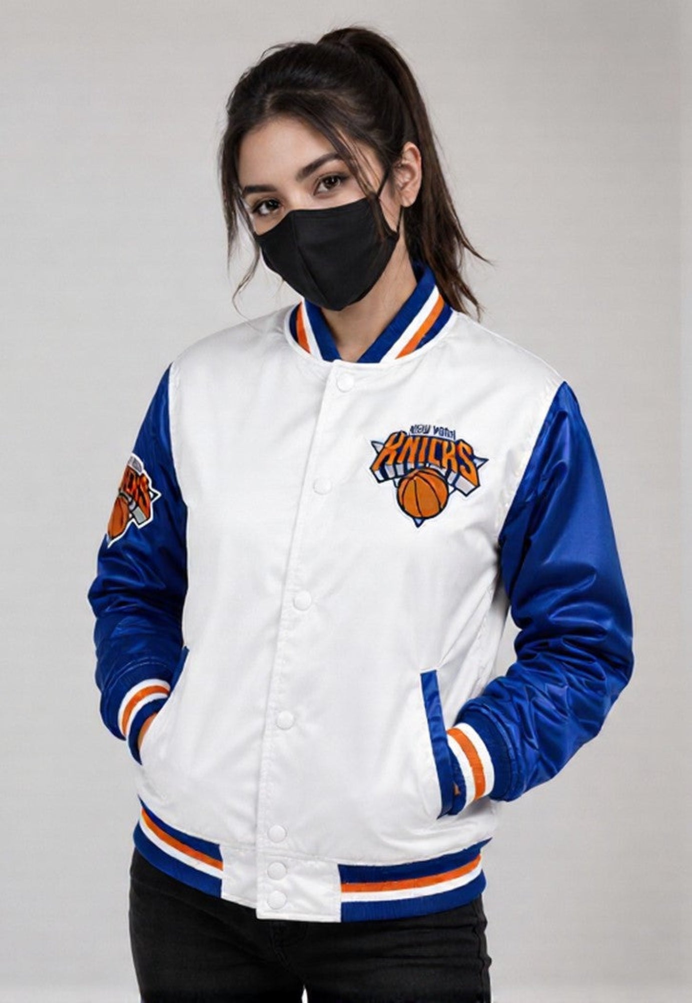 A woman wearing a white and blue New York Knicks varsity jacket with a black face mask, standing against a light gray studio background.