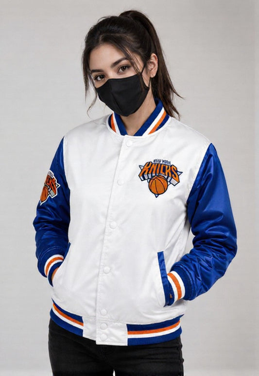 A woman wearing a white and blue New York Knicks varsity jacket with a black face mask, standing against a light gray studio background.