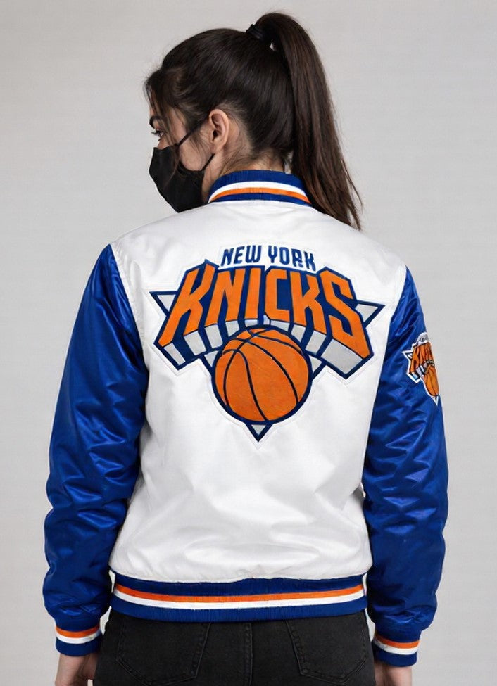 A woman shown from the back wearing a New York Knicks varsity-style jacket. The jacket has a white body, royal blue sleeves, and a large orange, blue, and white Knicks logo with a basketball graphic on the back.