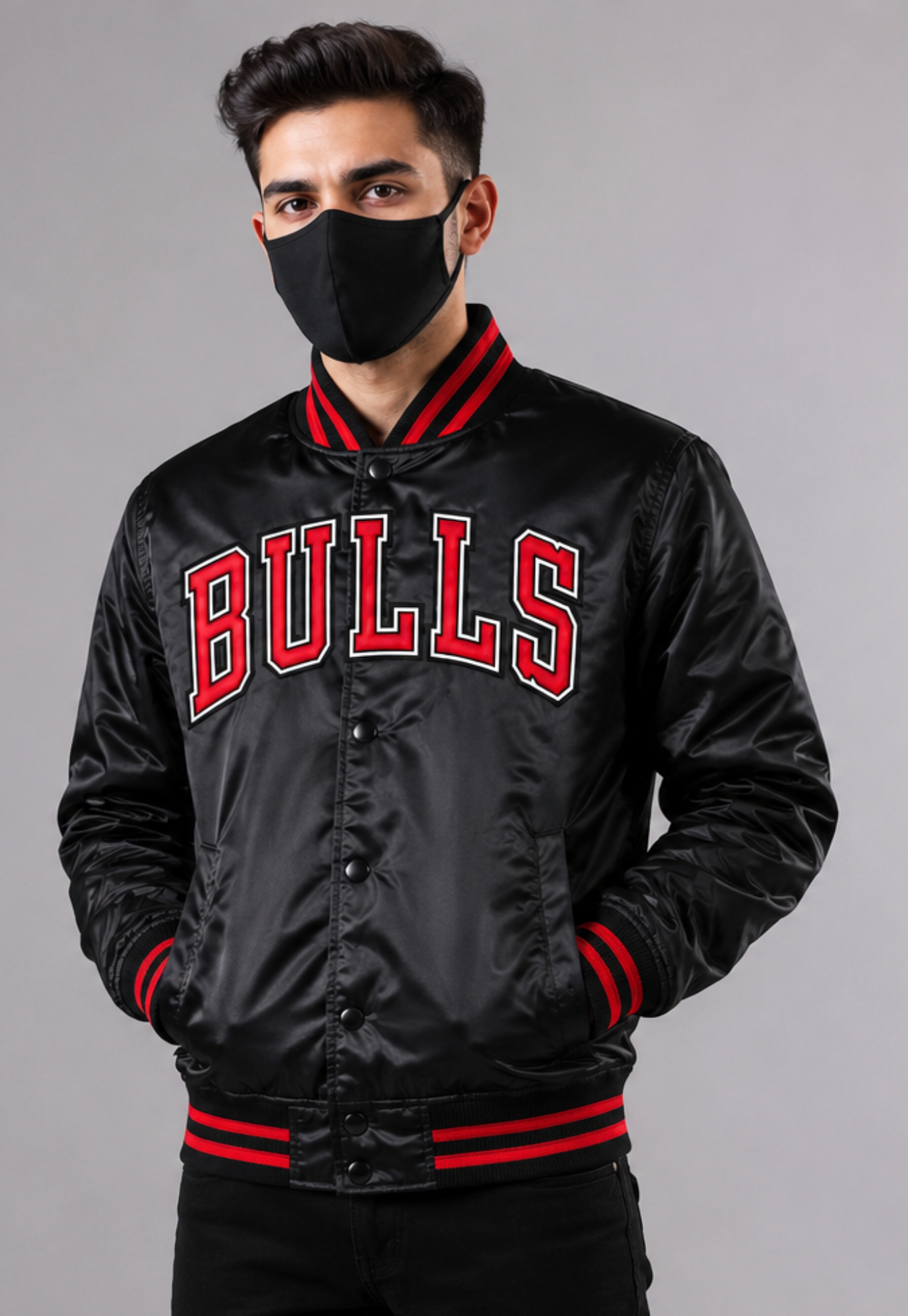 A man wearing a black satin varsity jacket with 'BULLS' in red and white on the chest, paired with a black face mask, against a solid gray studio background.