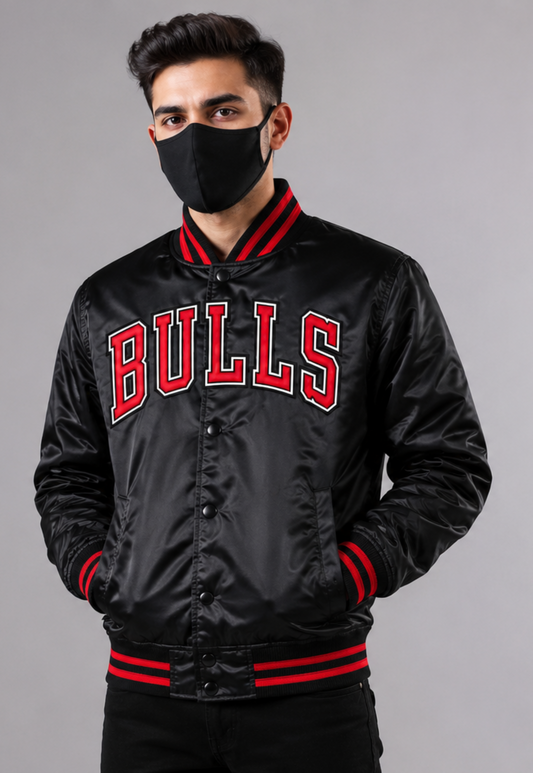 A man wearing a black satin varsity jacket with 'BULLS' in red and white on the chest, paired with a black face mask, against a solid gray studio background.