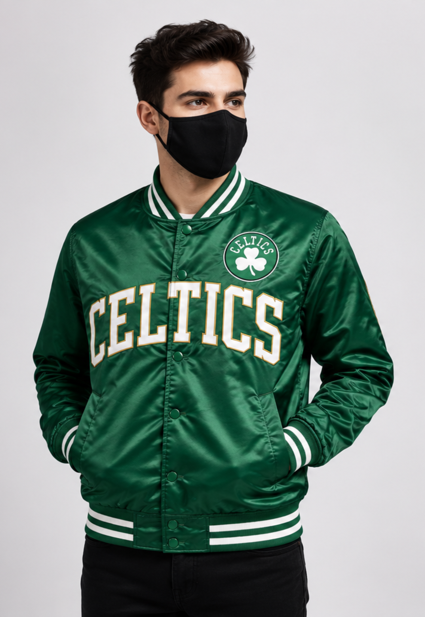 A man wearing a green Boston Celtics satin bomber jacket with white and green striped ribbed collar, cuffs, and hem, paired with a black face mask and black pants, against a light gray studio background.
