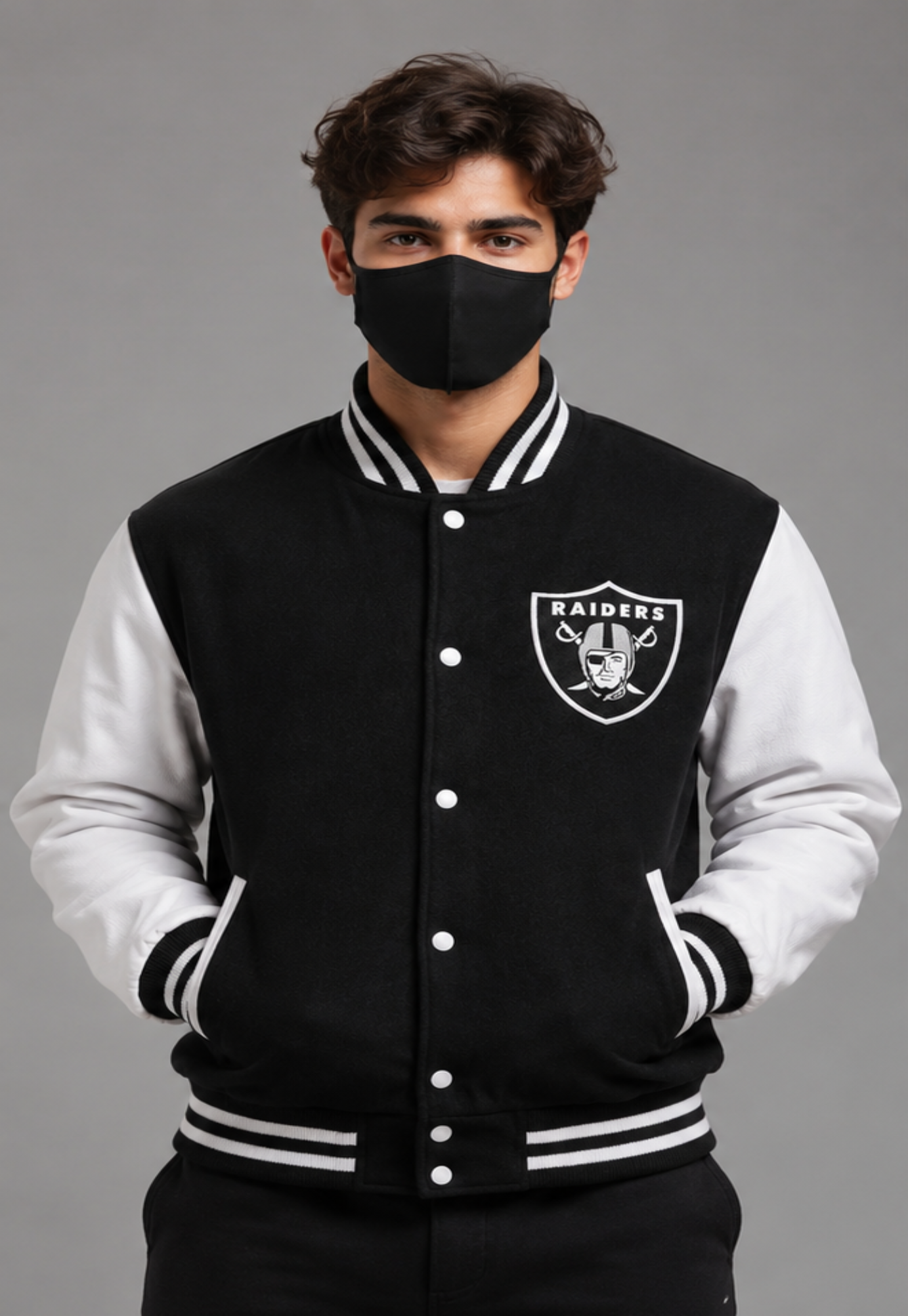A man wearing a black and white Oakland Raiders varsity jacket, featuring the team logo on the left chest, set against a solid gray studio background.