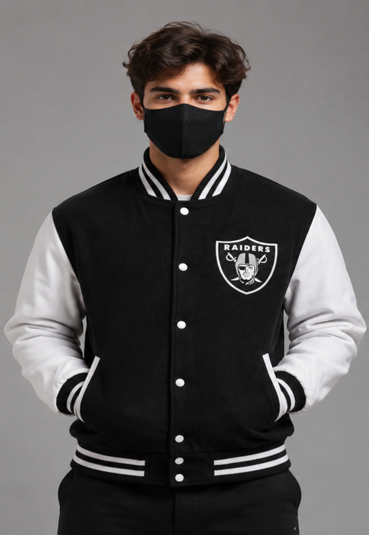 A man wearing a black and white Oakland Raiders varsity jacket, featuring the team logo on the left chest, set against a solid gray studio background.