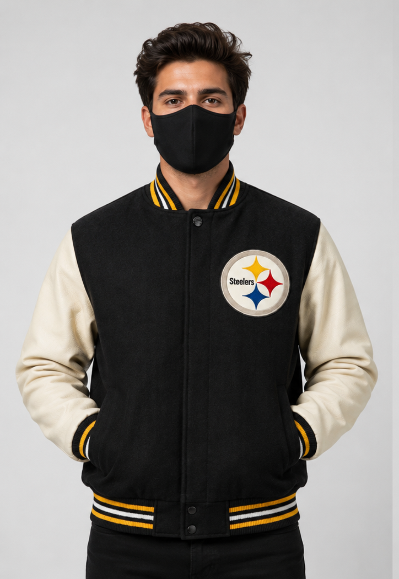 A man wearing a Pittsburgh Steelers varsity jacket with a black fleece body and cream sleeves, paired with a black face mask, against a light gray studio background.