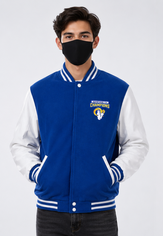 A man wearing a blue and white varsity jacket with a black face mask, standing against a light gray studio background. The jacket features a royal blue body with white sleeves and white striped ribbed trim, and a Los Angeles Rams logo on the left chest.