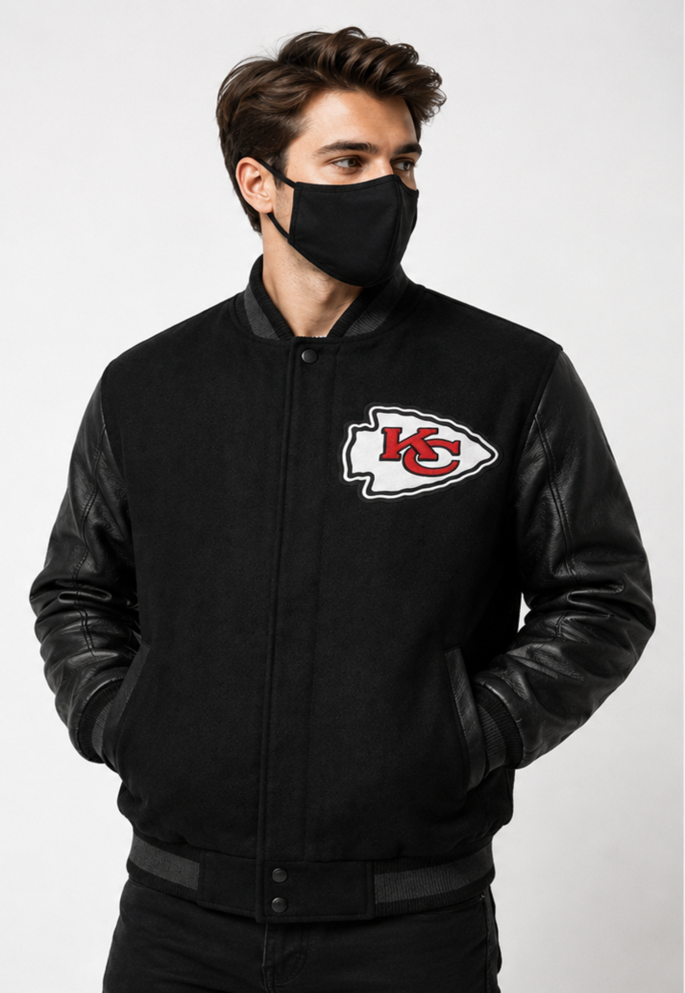 A man wearing a black Kansas City Chiefs varsity-style jacket with black leather sleeves and a black face mask, shown against a light gray studio background.