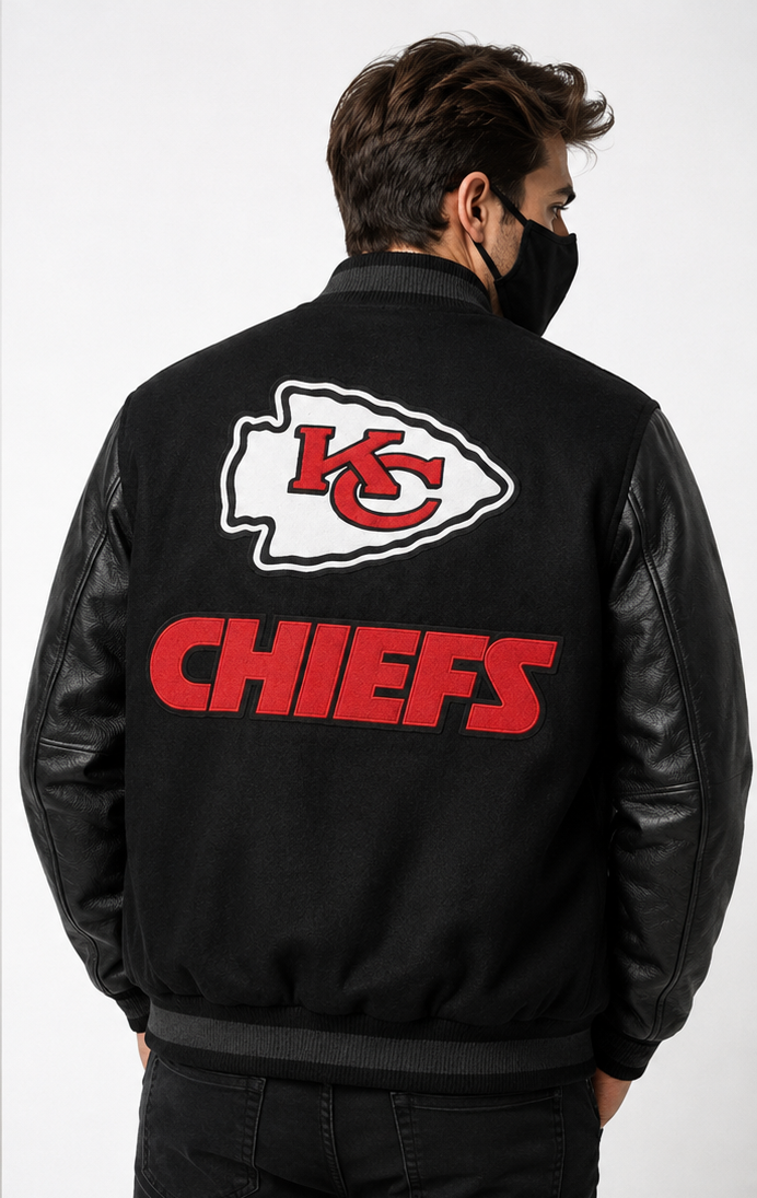 A man viewed from behind wearing a black leather-look bomber jacket with the Kansas City Chiefs logo and 'CHIEFS' text on the back, paired with a black face mask and dark pants, against a light gray studio background.