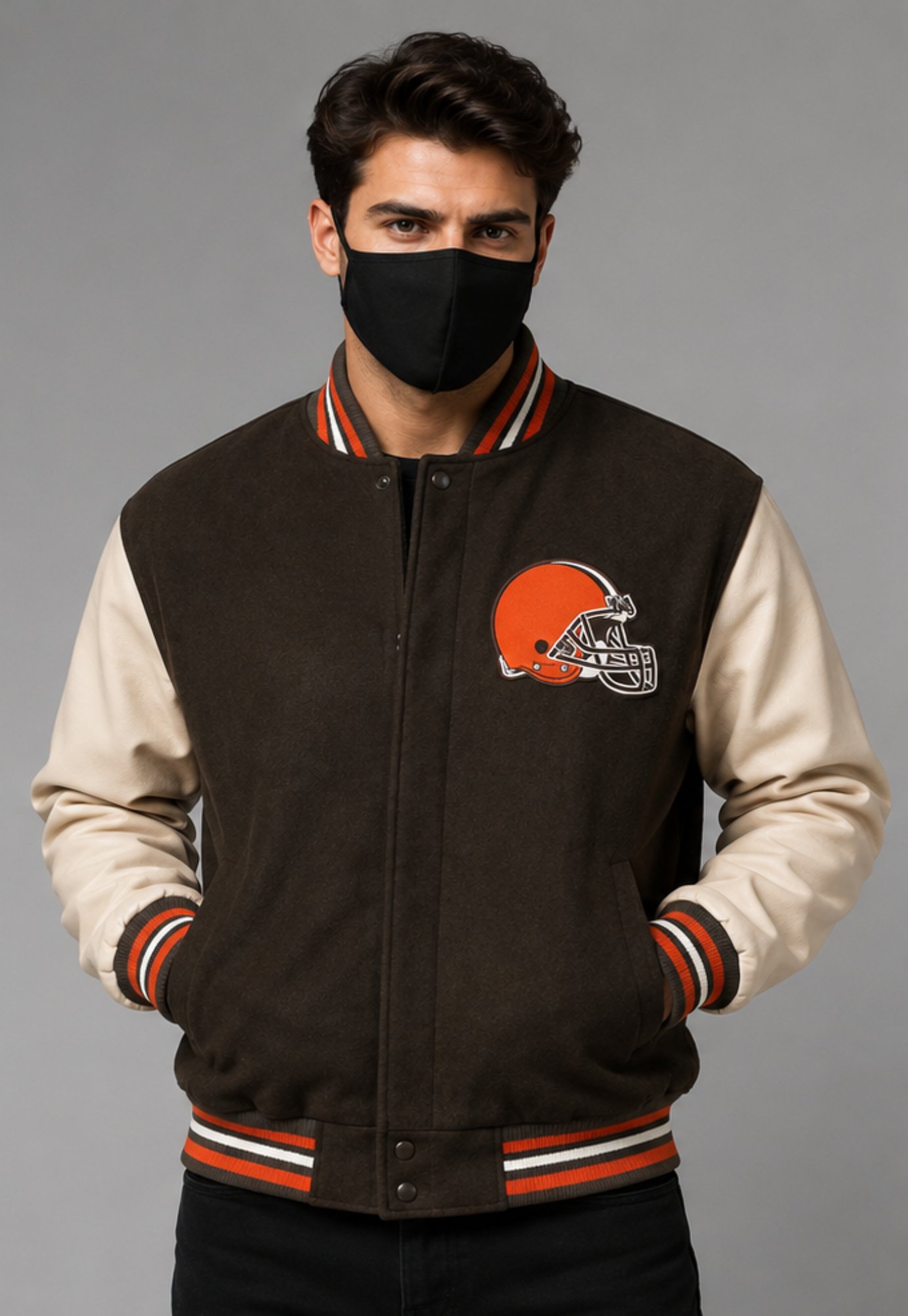 A man wearing a brown and cream varsity jacket with the Cleveland Browns helmet logo on the chest, paired with a black face mask, against a solid gray studio background.