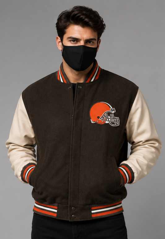 A man wearing a brown and cream varsity jacket with the Cleveland Browns helmet logo on the chest, paired with a black face mask, against a solid gray studio background.