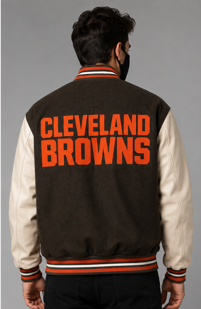 A man viewed from behind wearing a brown and cream varsity jacket with 'CLEVELAND BROWNS' embroidery in orange across the back, set against a plain gray studio background.
