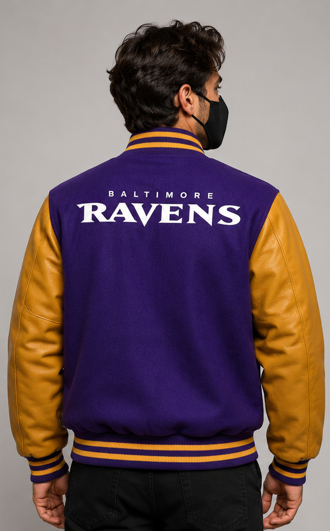A man viewed from the back wearing a Baltimore Ravens varsity jacket with a purple body, gold sleeves, and white lettering on the back, photographed against a plain light gray studio background.