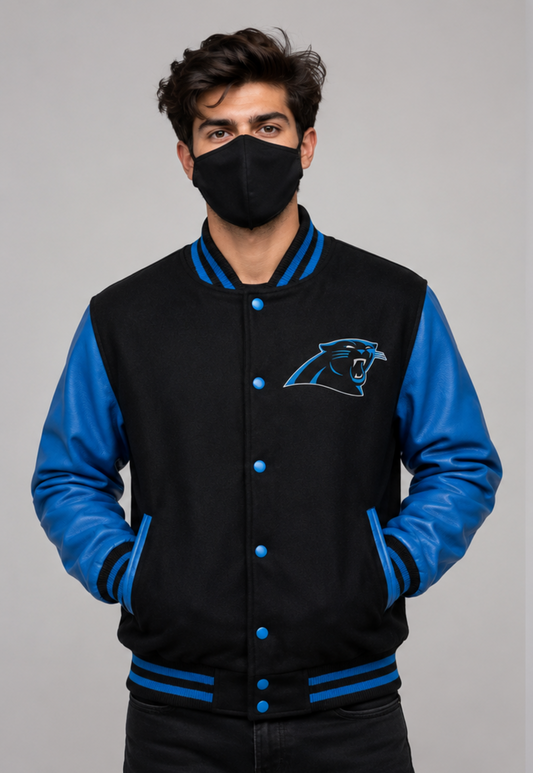 A man wearing a black and blue varsity jacket with a Carolina Panthers logo on the chest, paired with a black face mask, against a solid gray background.