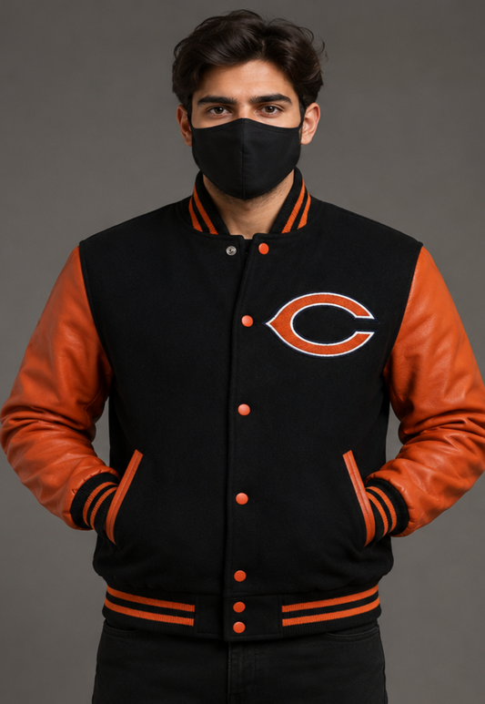 A man wearing a black varsity jacket with orange leather sleeves and a large Chicago Bears logo on the chest, paired with a black face mask, against a solid gray studio background.